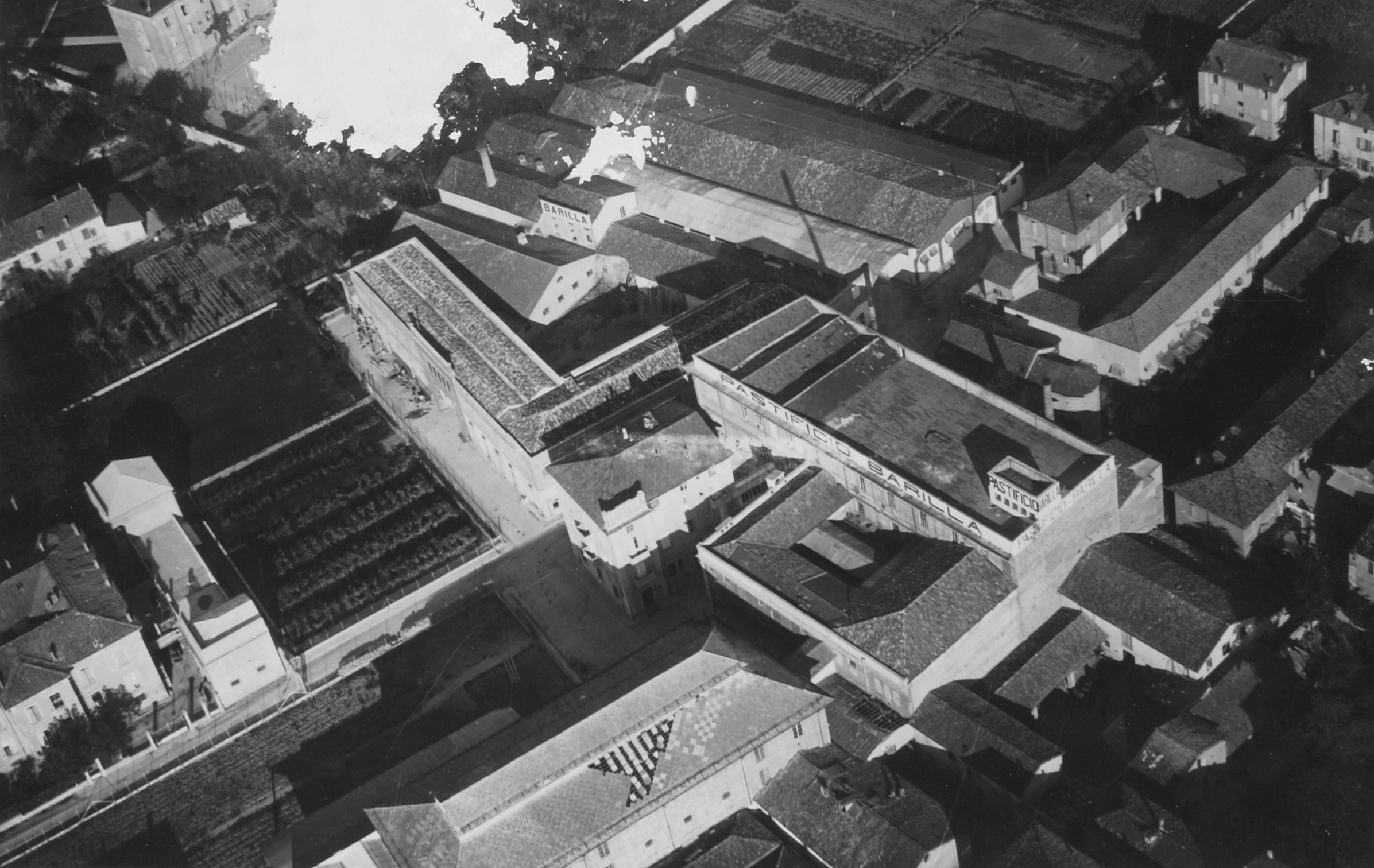 Una vista dall’alto, ripresa da Sud-Ovest dalla NAIS di Siena, successiva il 1930 [ASB, A210]. Si possono riconoscere il pastificio, al centro, ingranditosi intorno ad un piccolo cortile centrale, successivamente coperto, il panificio dalla facciata in muratura a vista, dalla caratteristica pianta a forma di “L” e, in basso, l’edificio del “Mulino”, adibito a magazzini, con il tetto in rifacimento.